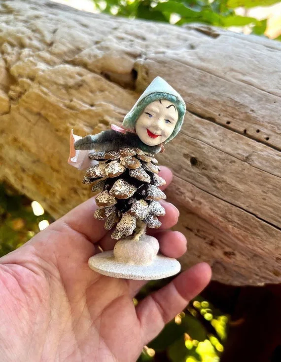 A hand holds a small, handmade figurine of a smiling girl with a green hood, whose body is crafted from a pinecone and…