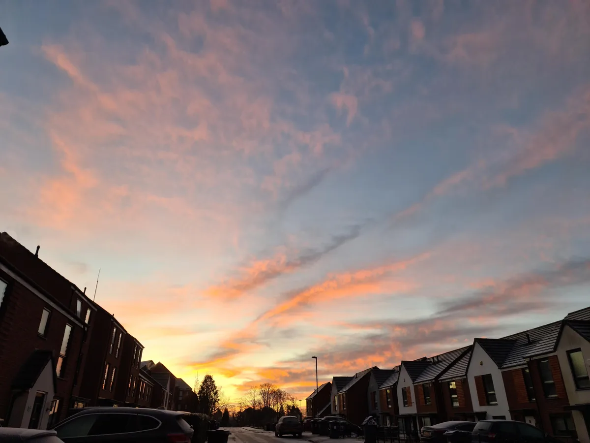 A photograph of a residential street at sunset, with rows of houses on both sides and a sky filled with pink, orange, and…
