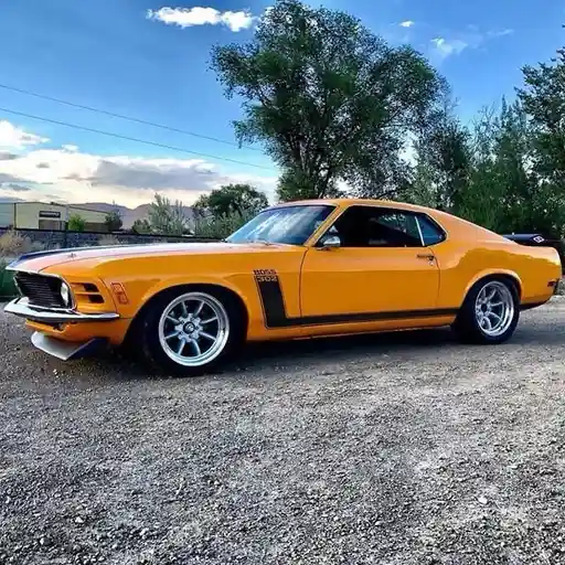 a bright orange 1970 ford mustang boss 429 is parked on gravel under a partly cloudy blue sky, with trees and distant hills…