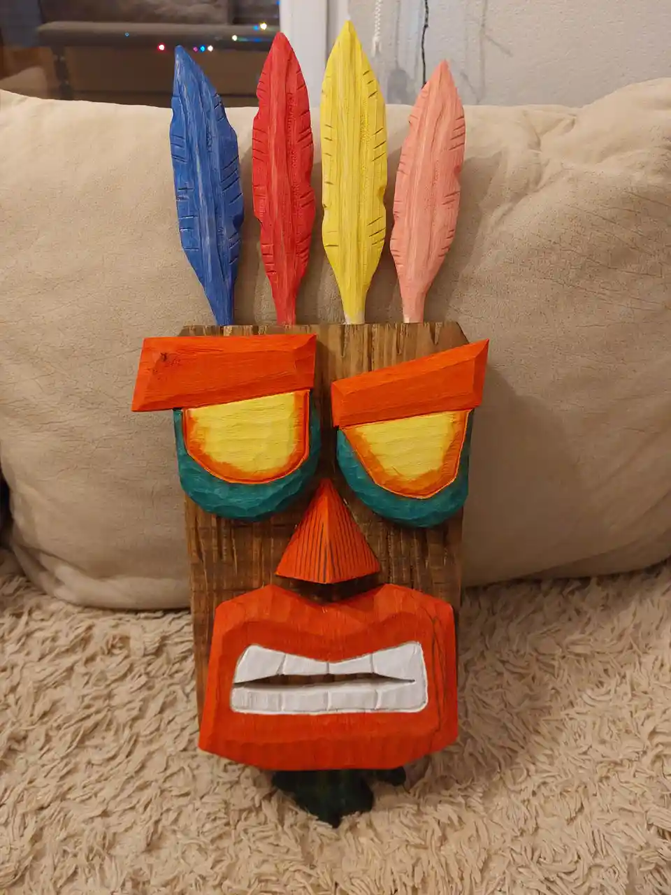A hand-carved wooden tiki mask with four colorful feathers (blue, red, yellow, pink) atop its head sits on a shaggy beige…
