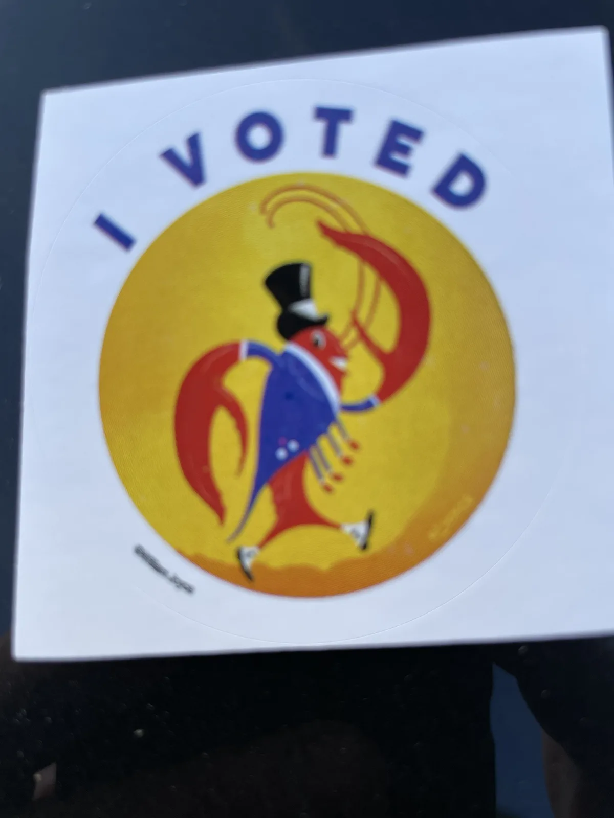 A circular 'I VOTED' sticker features a stylized red lobster wearing a black top hat and blue vest, dancing against a yellow…