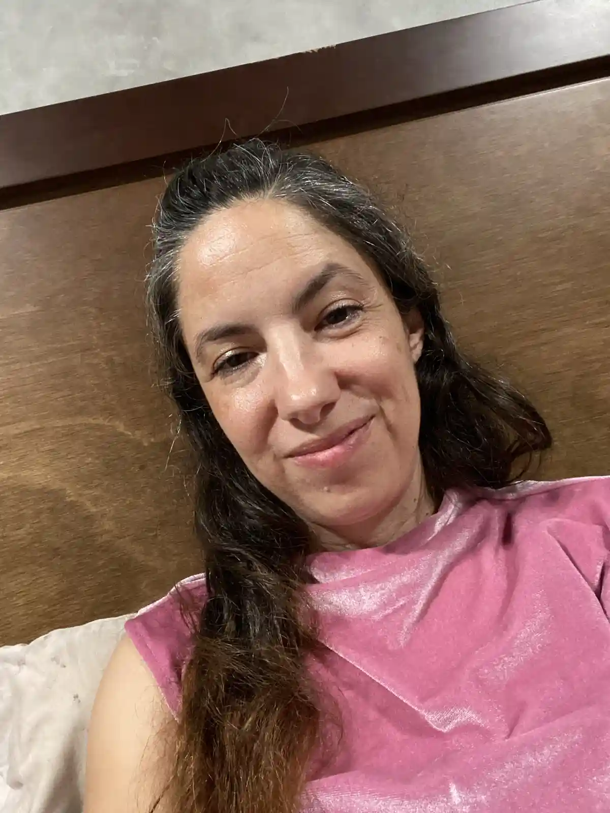 A woman with dark hair showing gray roots and a subtle smile is lying down, wearing a pink salon cape, against a wooden…