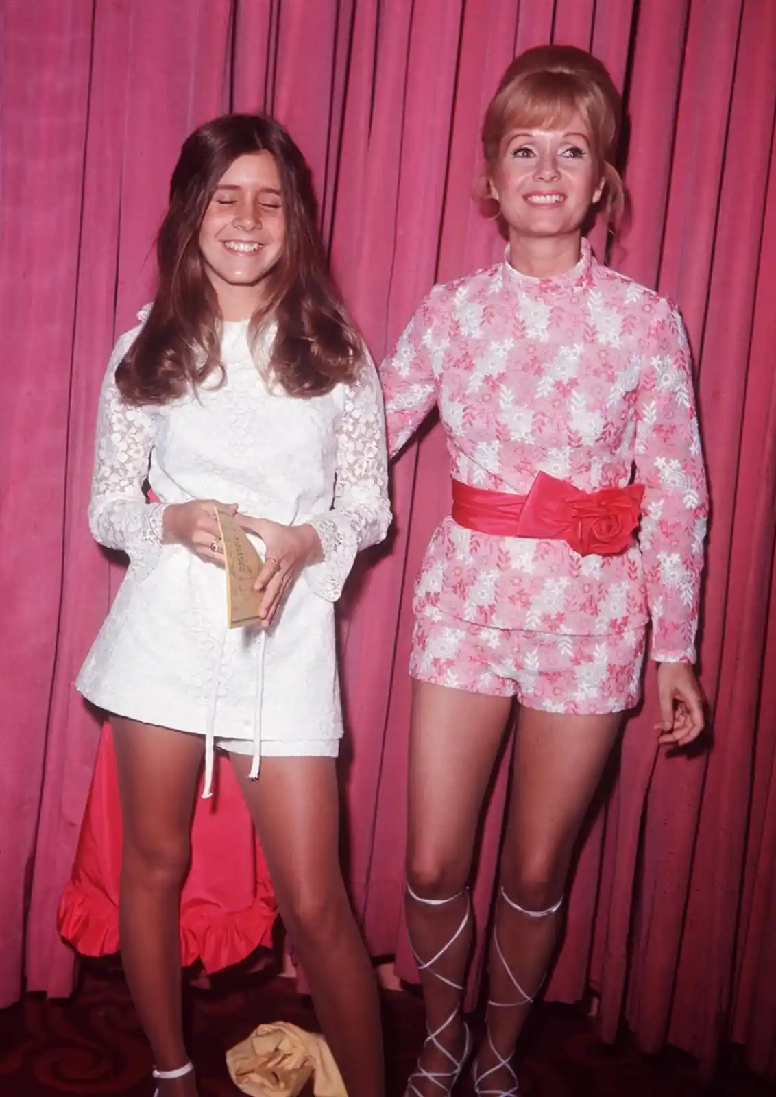Two women pose in front of a pink curtain; the woman on the left wears a white lace dress and smiles with her eyes closed…