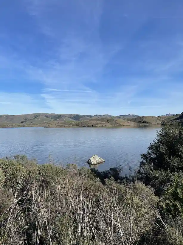 A serene landscape photo shows a calm lake under a clear blue sky, with rolling hills in the distance and a large rock…