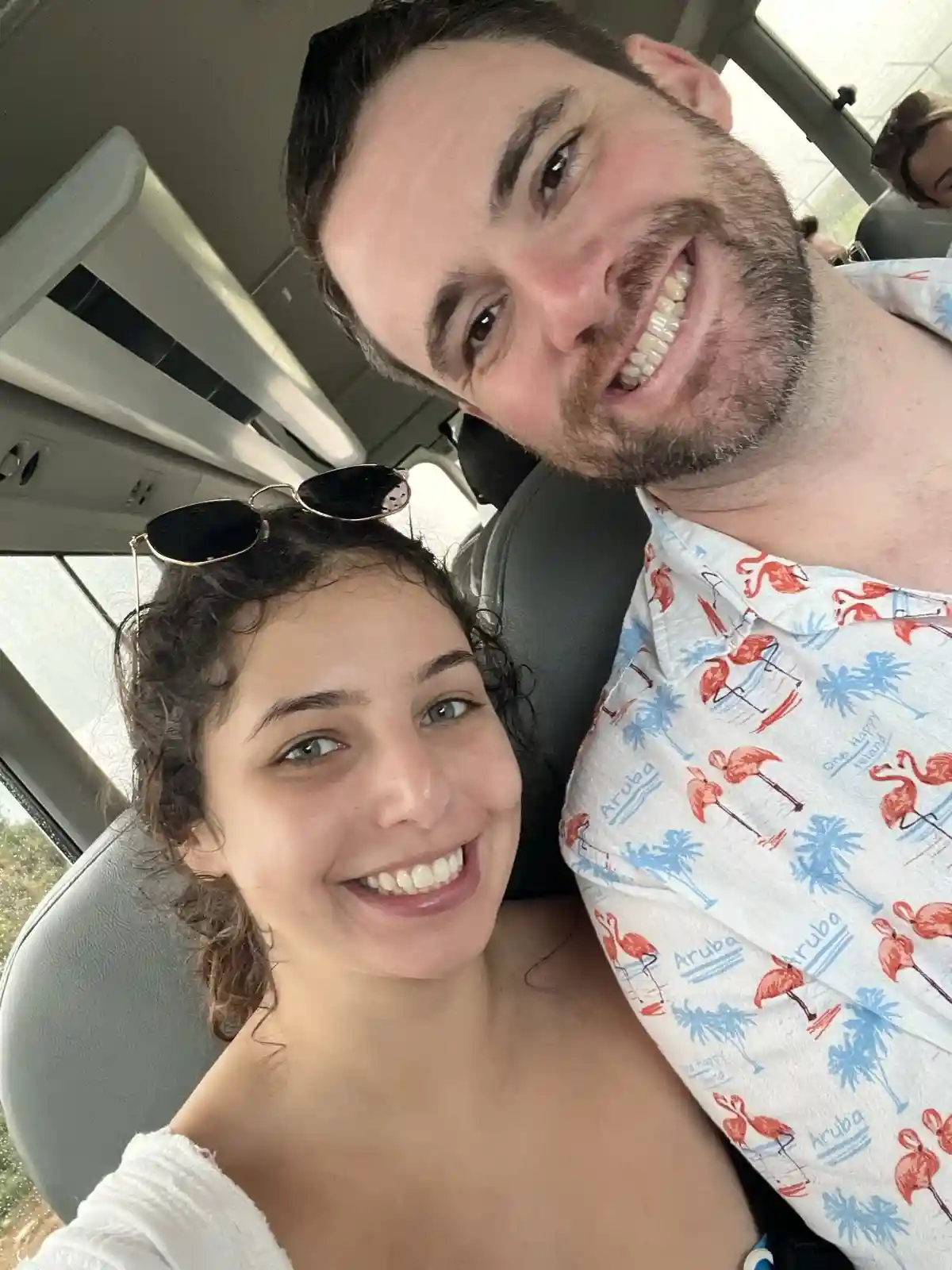 A selfie of a smiling man and woman inside a vehicle, the man wearing a white shirt with red flamingos and blue palm trees…