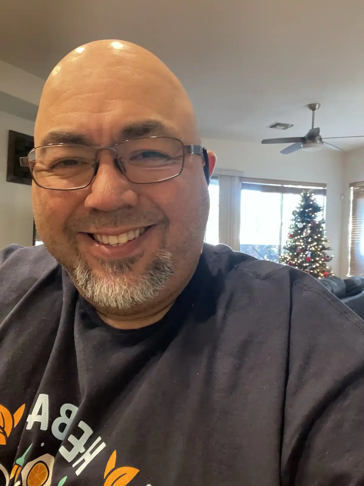 A bald man with glasses and a goatee smiles at the camera while wearing a dark t-shirt with a partial graphic. In the…