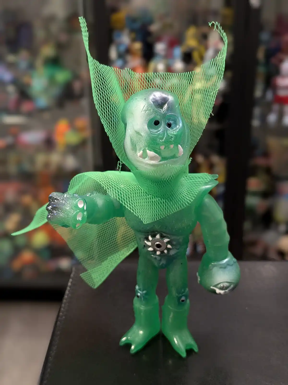 A translucent green plastic monster toy with a mesh cape and multiple eyes is posed on a black surface, with a blurred…
