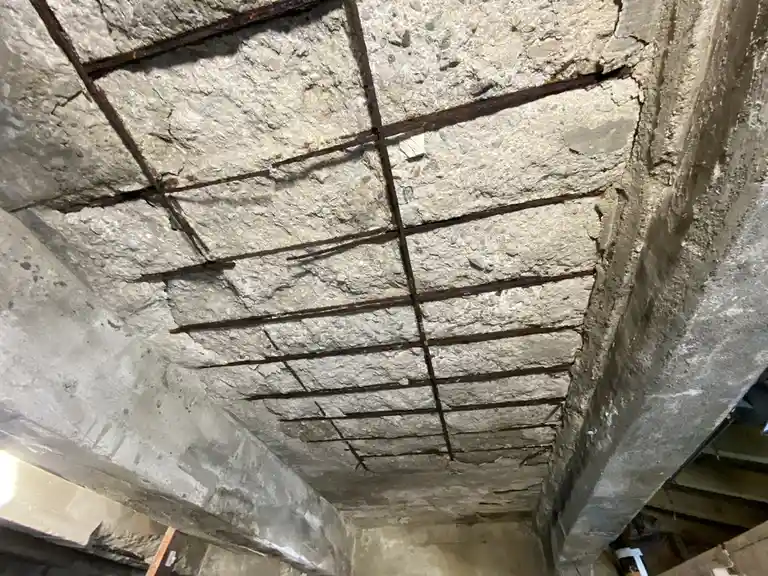 A close-up, upward-looking shot of a damaged concrete ceiling with exposed, rusted rebar forming a grid pattern. The…