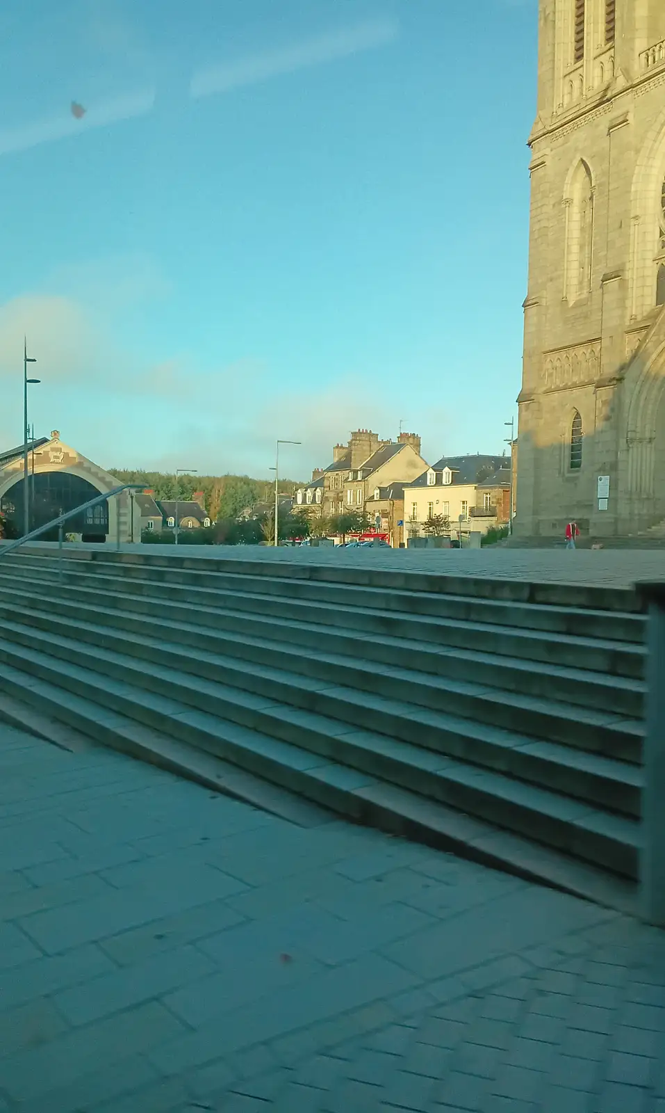 A low-angle photo shows wide concrete steps leading up to a plaza with a large stone church tower on the right and other…