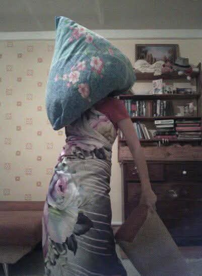 A person viewed from behind, wearing a floral dress, has a large floral-patterned pillow balanced on their head. They are…