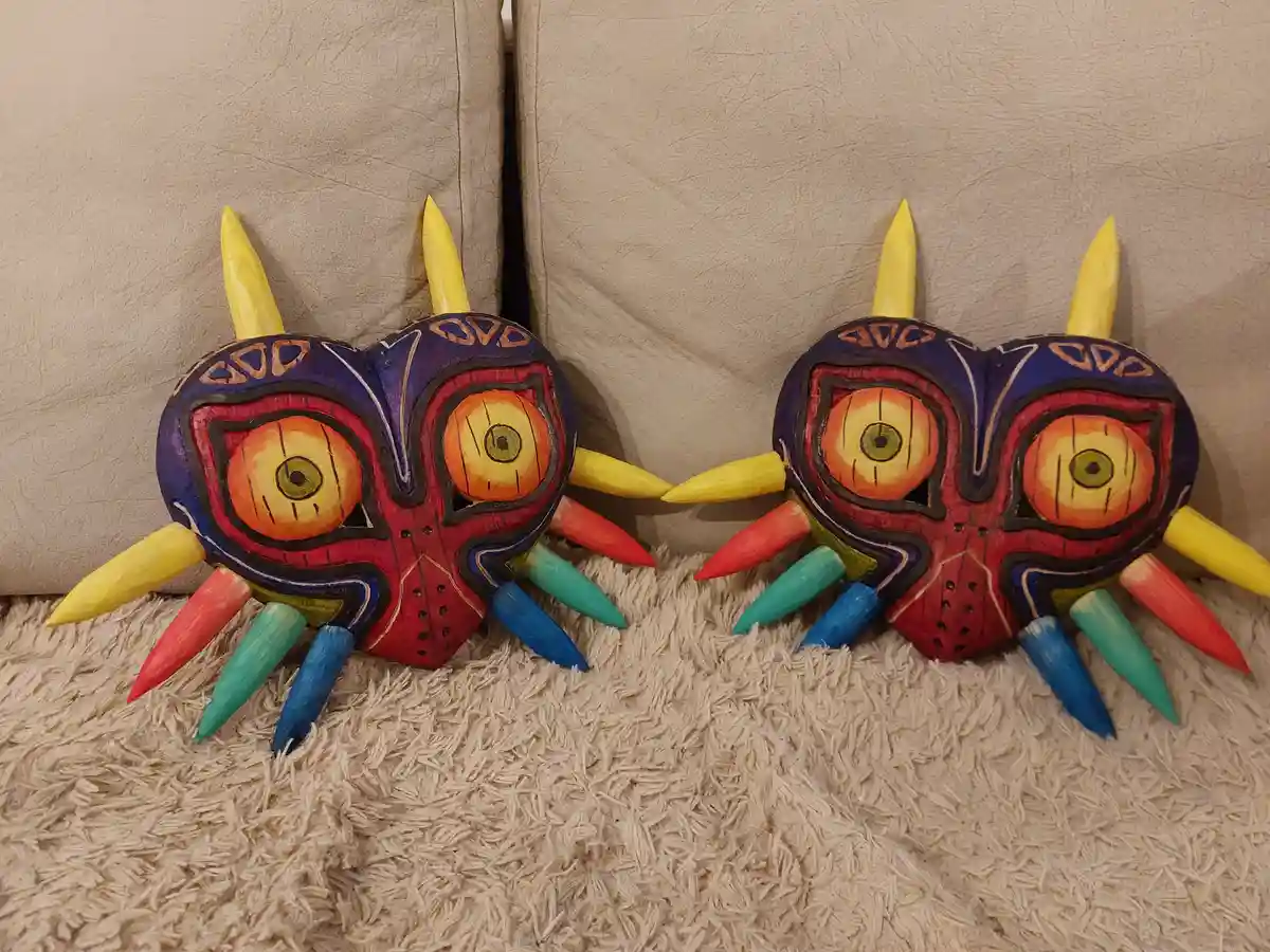 Two identical, colorful, hand-painted wooden masks resembling Majora's Mask from The Legend of Zelda, with red, blue, and…