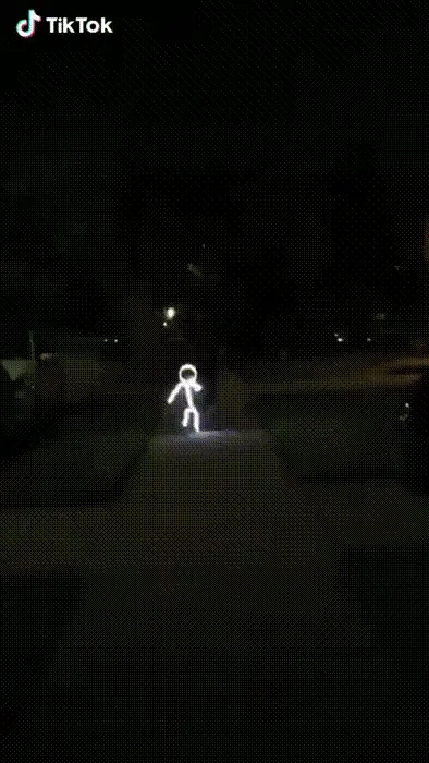 A person wearing a glowing stick-figure outline costume walks down a dark sidewalk at night, then the costume's headpiece…