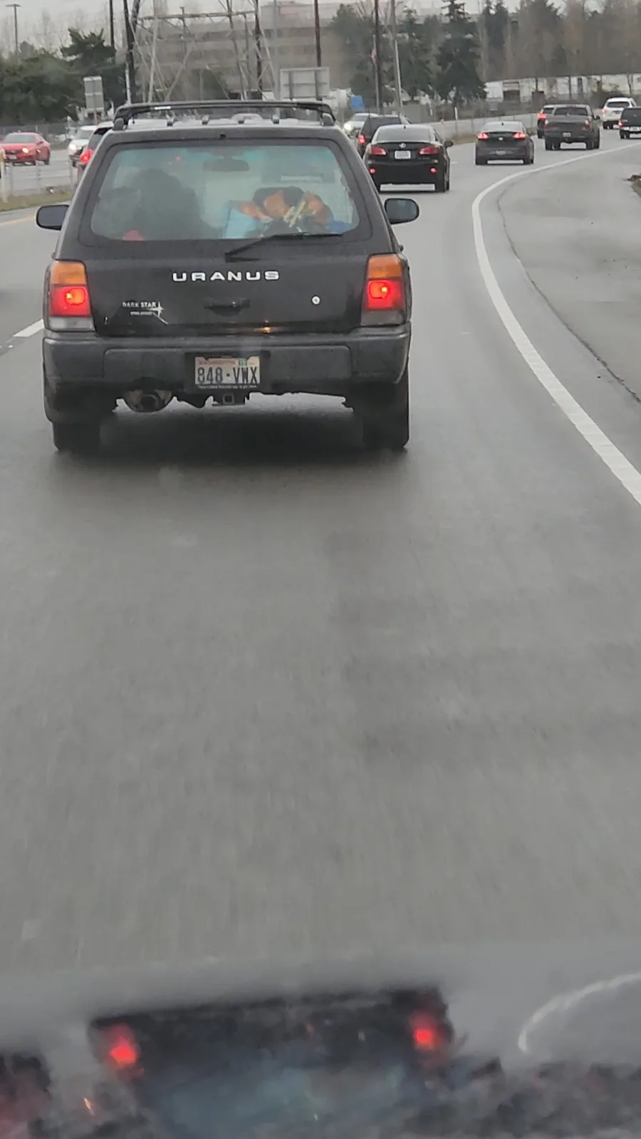 A black car with 'URANUS' written on the rear hatch is seen from behind while driving on a highway. Through the rear window…