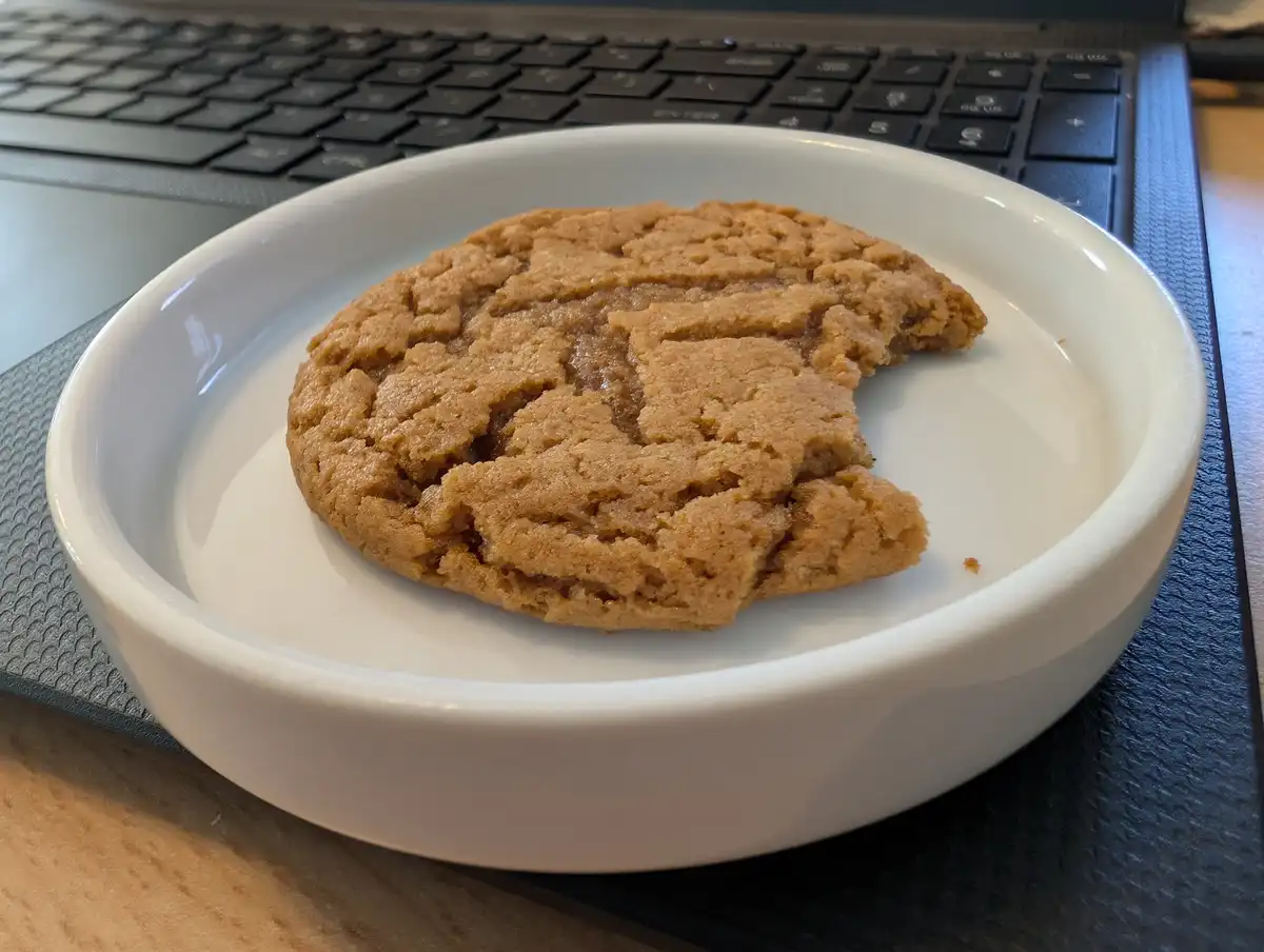 A partially eaten peanut butter cookie sits on a white plate, positioned in front of a laptop keyboard. The cookie has a…