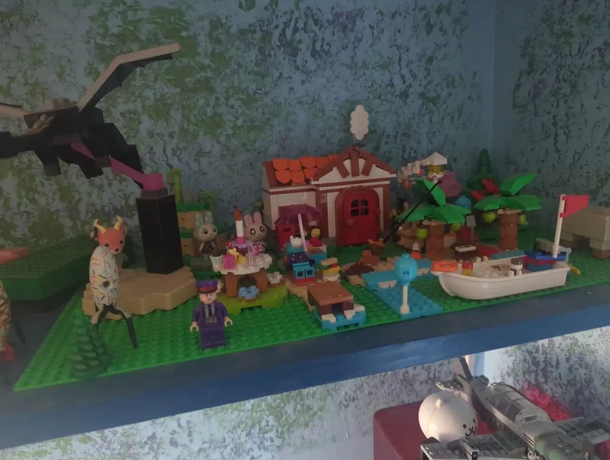 A colorful Lego diorama depicts a whimsical village scene with a house, palm trees, a boat, and various animal and human…