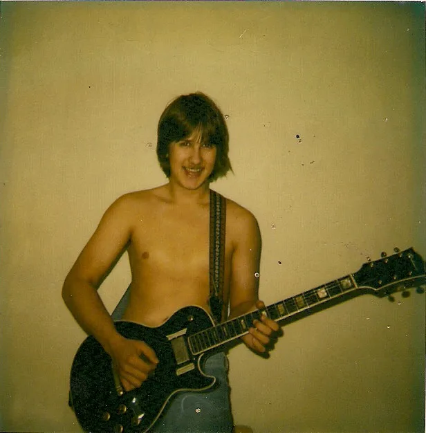 A grainy, vintage photograph shows a young, shirtless man with shaggy hair smiling while holding a black electric guitar…