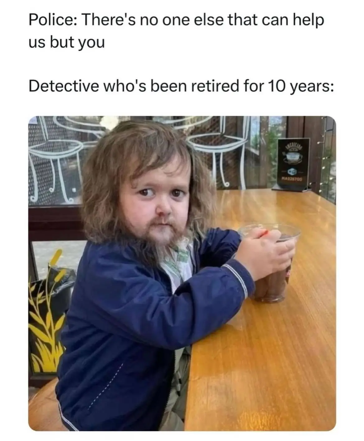 A young child with long brown hair and a beard sits at a wooden table in a cafe, holding a plastic cup, looking unimpressed…