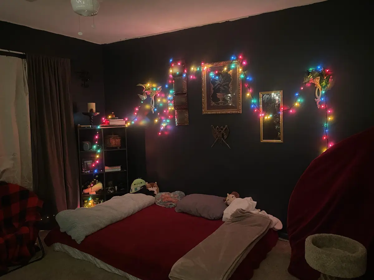A dimly lit bedroom with dark walls, a bed covered in red and grey blankets, and colorful string lights arranged in a…
