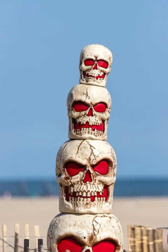 A stack of four weathered, white plastic skulls with glowing red eyes is arranged vertically on a beach, with a clear blue…