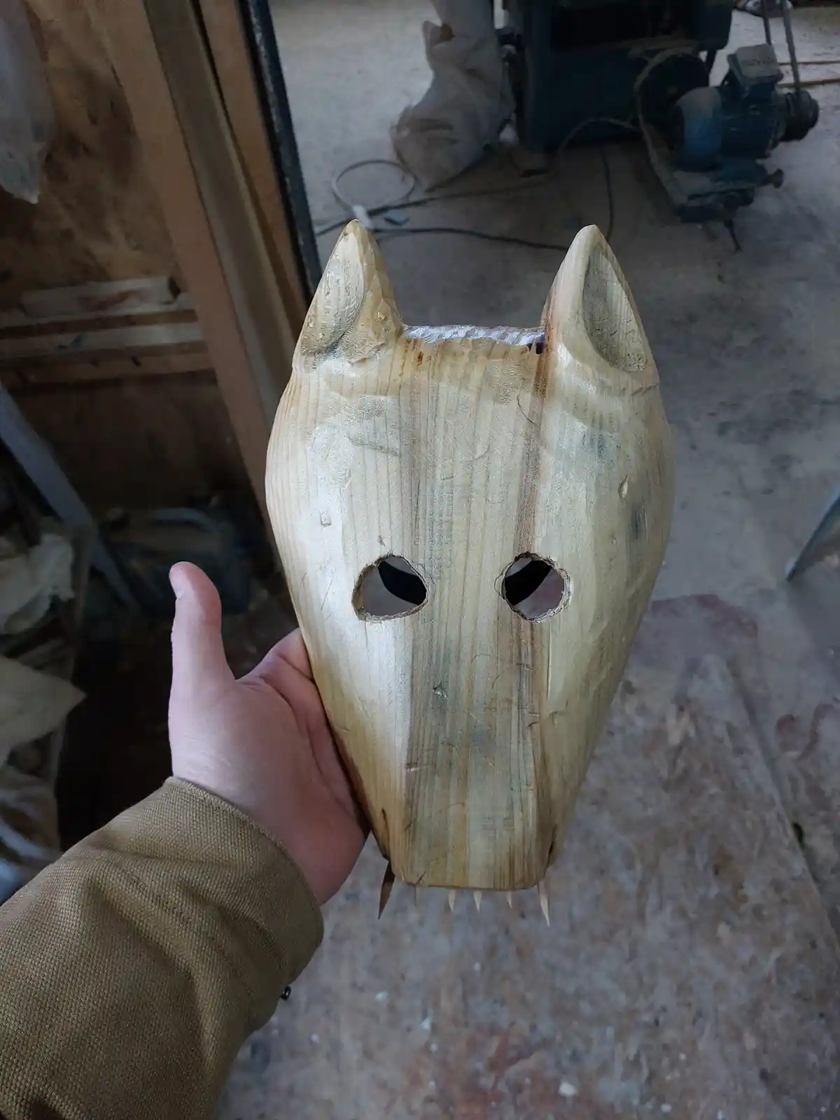 A person holds up a crudely carved wooden mask shaped like a wolf or dog, featuring pointed ears, two rough eye holes, and…
