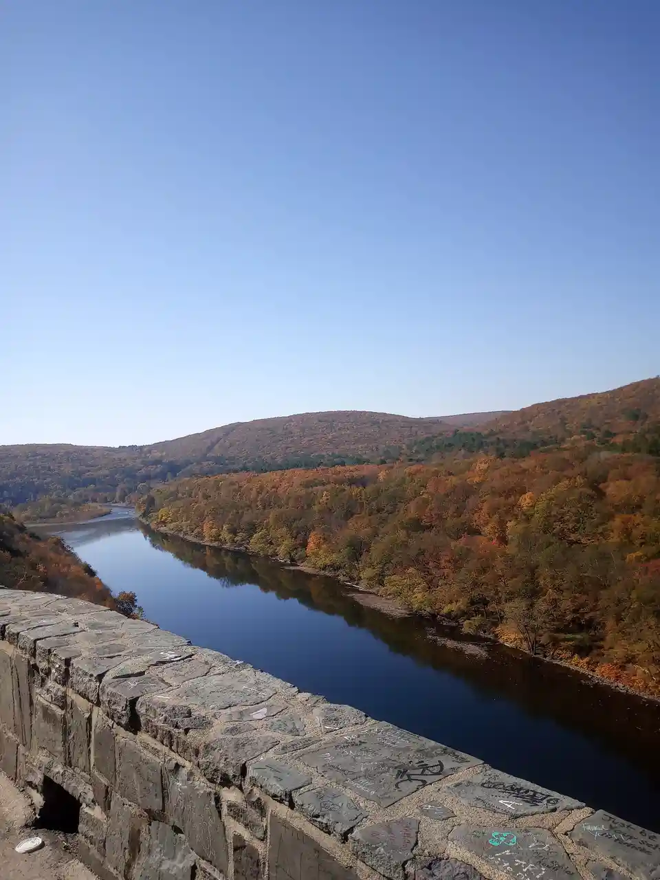 A scenic autumn view of a river winding through forested hills under a clear blue sky, framed by a stone wall in the…