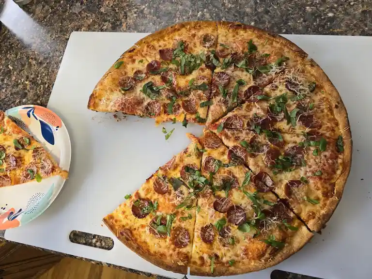 A freshly baked pepperoni pizza with melted cheese and basil leaves sits on a white cutting board, with one slice removed…