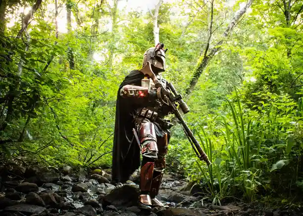 A person in full Mandalorian armor, complete with helmet, cape, and blaster rifle, stands in a lush green forest on a rocky…