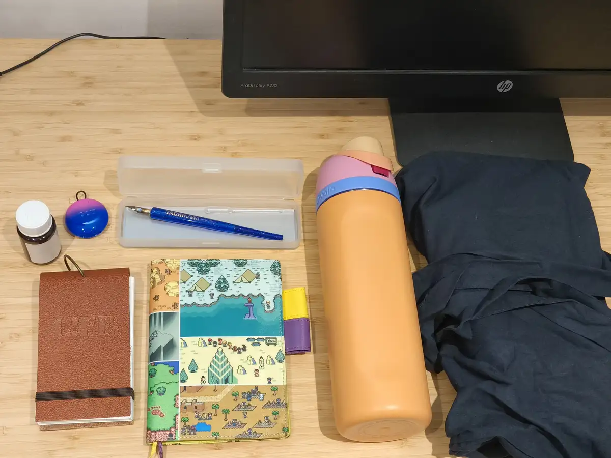 A wooden desk displays a neatly arranged collection of personal items including a brown leather notebook, a colorful…
