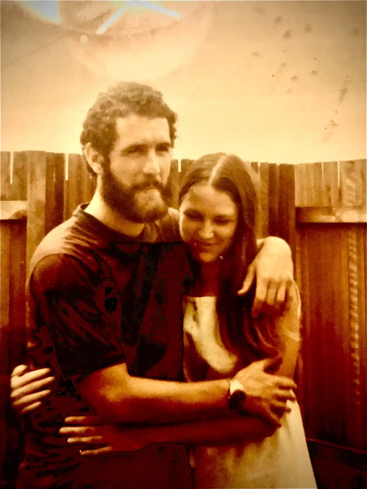 A sepia-toned, slightly blurry vintage photo shows a man with a beard and a woman with long hair embracing in front of a…