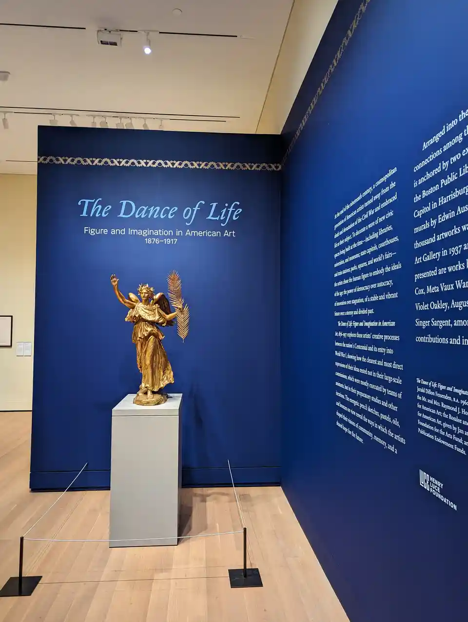 A golden statue of an angelic figure with wings, holding a palm frond and raising one hand, stands on a pedestal in a museum…