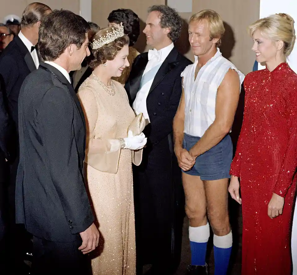 A formal royal event photo shows Queen Elizabeth II in a tiara and gown smiling while conversing with a man in a tuxedo…