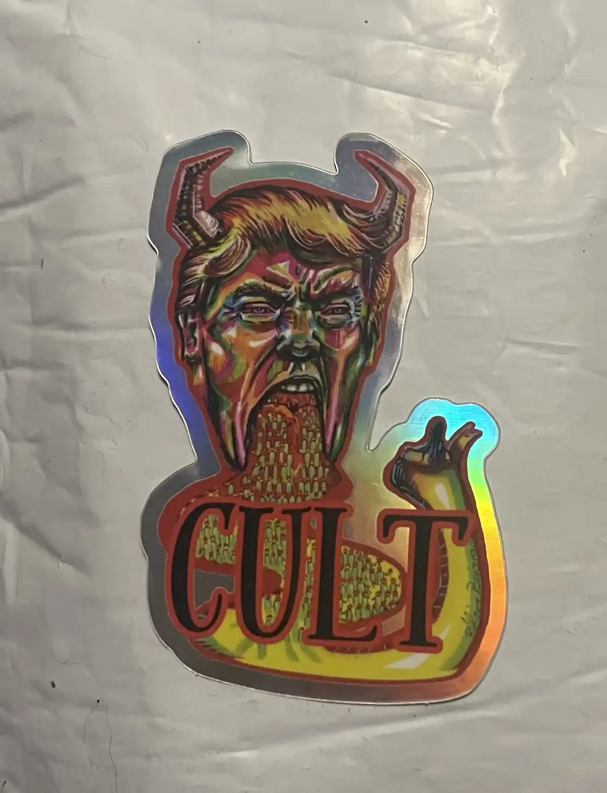 A holographic sticker depicts a demonic figure with horns and a screaming face, resembling Donald Trump, with the word…