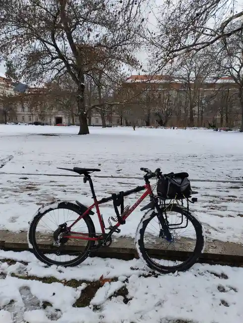 A red bicycle with black tires and a black bag strapped to its rear rack is parked on a snow-covered sidewalk in a park…
