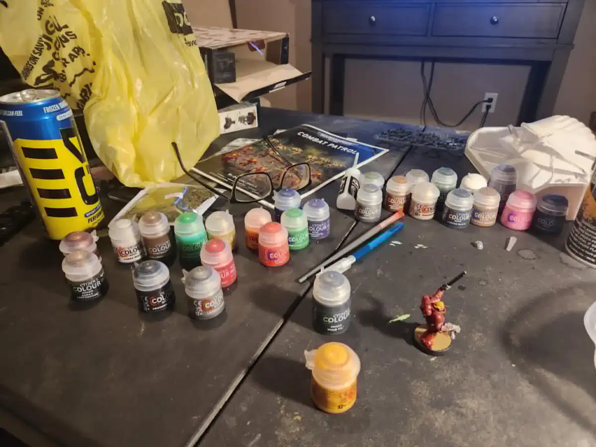 A cluttered tabletop is covered with numerous small pots of Citadel Colour paints, paintbrushes, a partially painted…