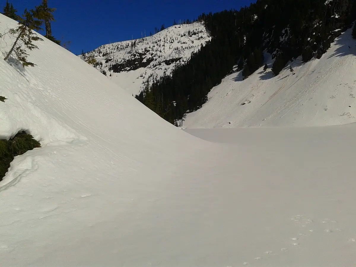 A snowy mountain valley with a wide, flat snowfield in the foreground and steep, snow-covered slopes rising on either side…