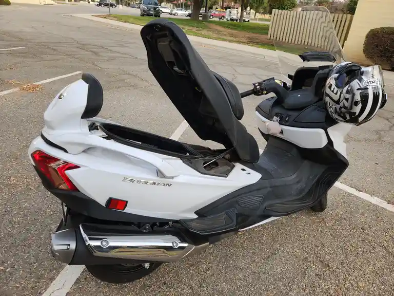 A white and black Honda Forza scooter is parked in a lot with its seat lifted up, revealing the storage compartment…