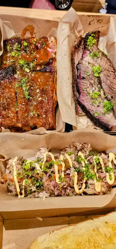 Three cardboard boxes display different types of cooked meat: ribs glazed with sauce, sliced brisket with a pink center, and…