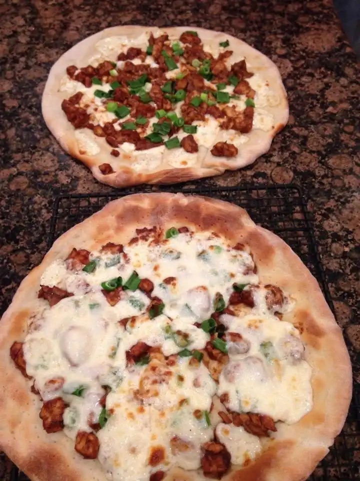 Two flatbreads sit on a countertop, one above the other. The top one has toppings and green onions but no melted cheese. The…