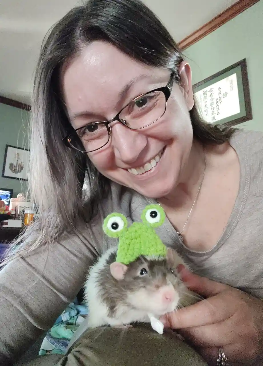 A woman with glasses smiles while holding a hamster wearing a green crocheted frog hat with large eyes. The hamster looks…