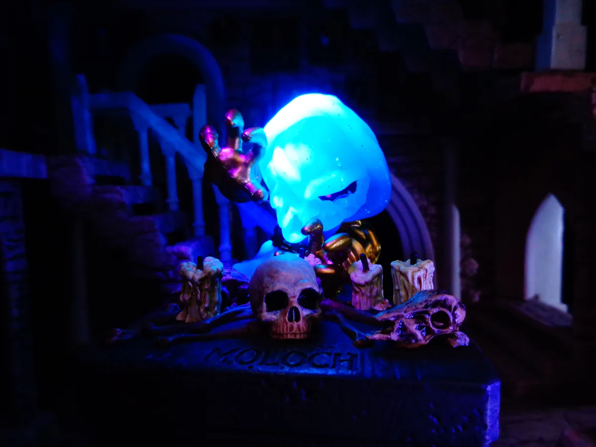 A glowing blue skull figurine with a golden hand raised, sits atop a dark stone altar engraved with 'MOLOCH', surrounded by…