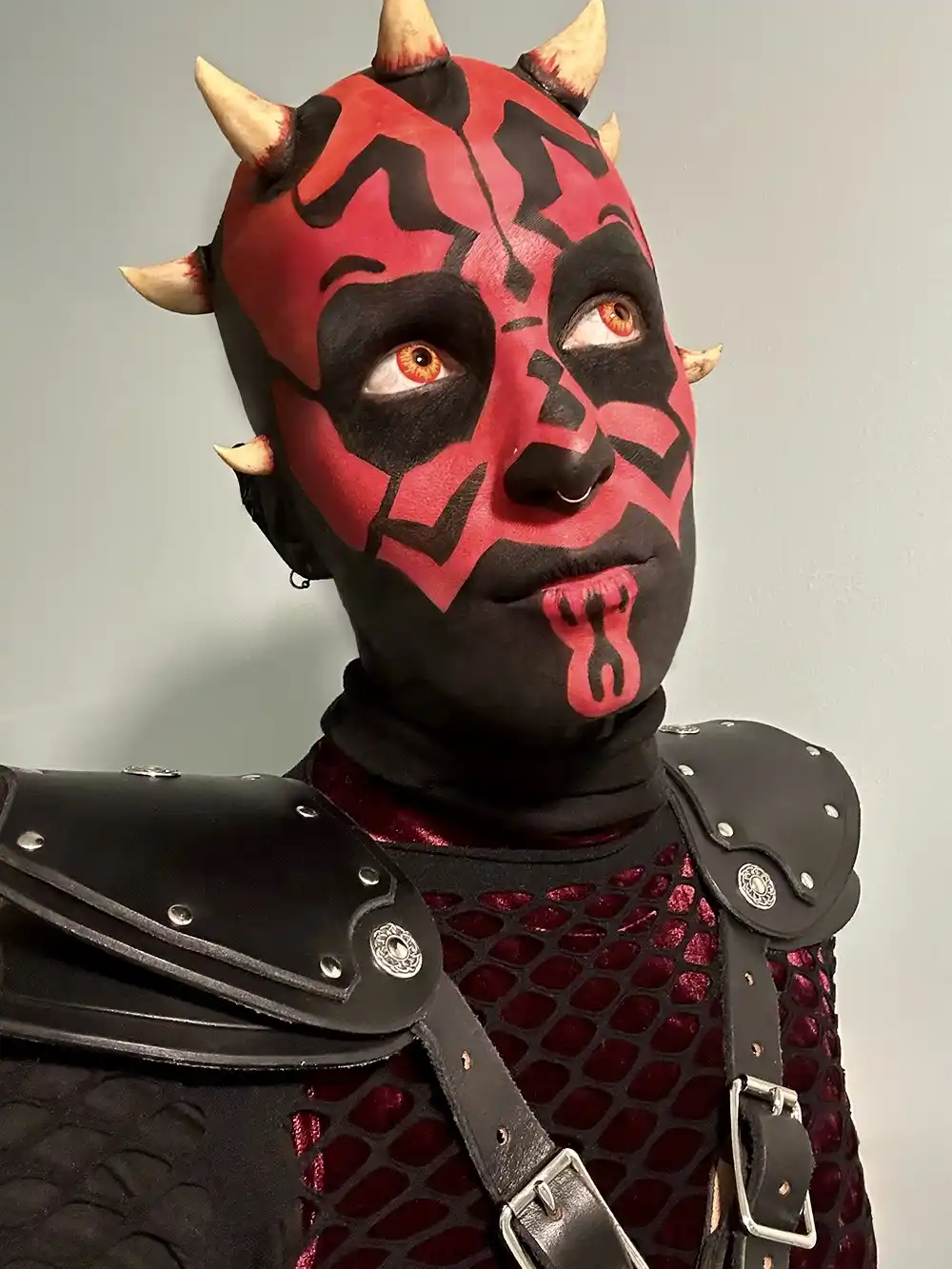 A person in full Darth Maul cosplay, with red and black face paint, horns, and dark armor, stares upward with a slightly…