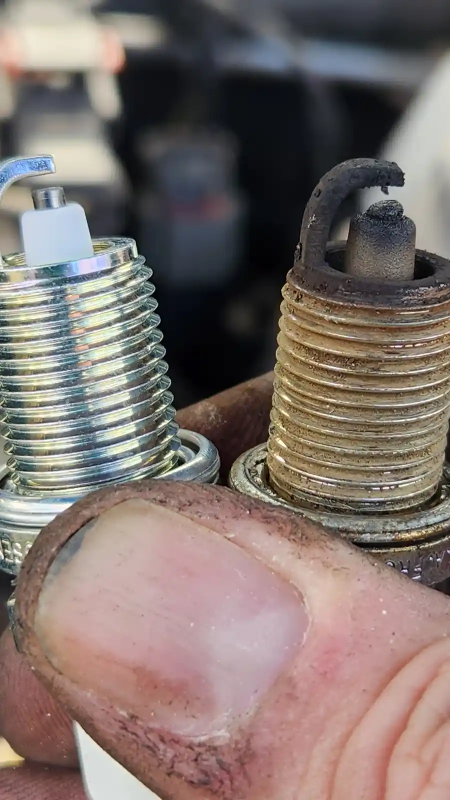 A close-up photo shows a hand holding two spark plugs side-by-side: one brand new and shiny with a white ceramic insulator…