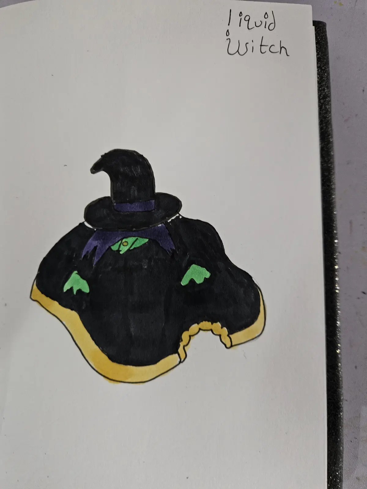 A hand-drawn illustration on white paper depicts a witch's hat and cloak shaped like a puddle or blob, with a yellow outline…