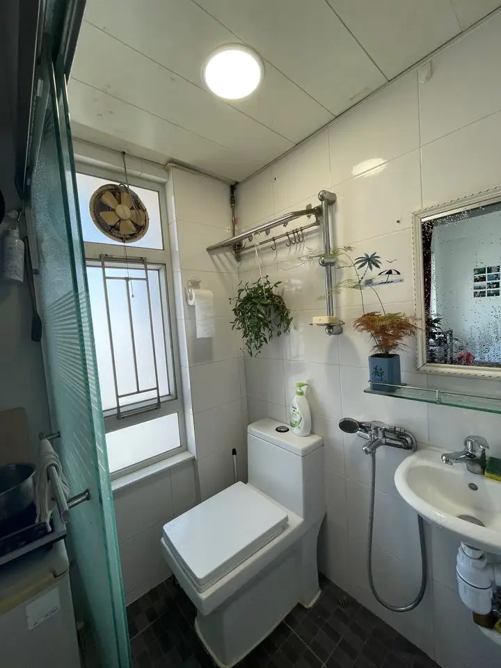 A small, white-tiled bathroom contains a toilet, sink, and shower area with a glass door. A window with a hanging fan is…