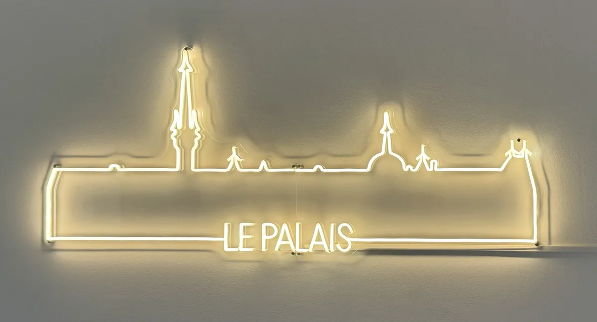 A glowing yellow neon sign mounted on a plain wall depicts the silhouette of a palace with multiple spires and turrets, and…