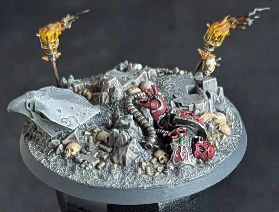 A detailed miniature wargaming base depicts a fallen, red-armored warrior amidst rubble, skulls, and debris. Two flaming…