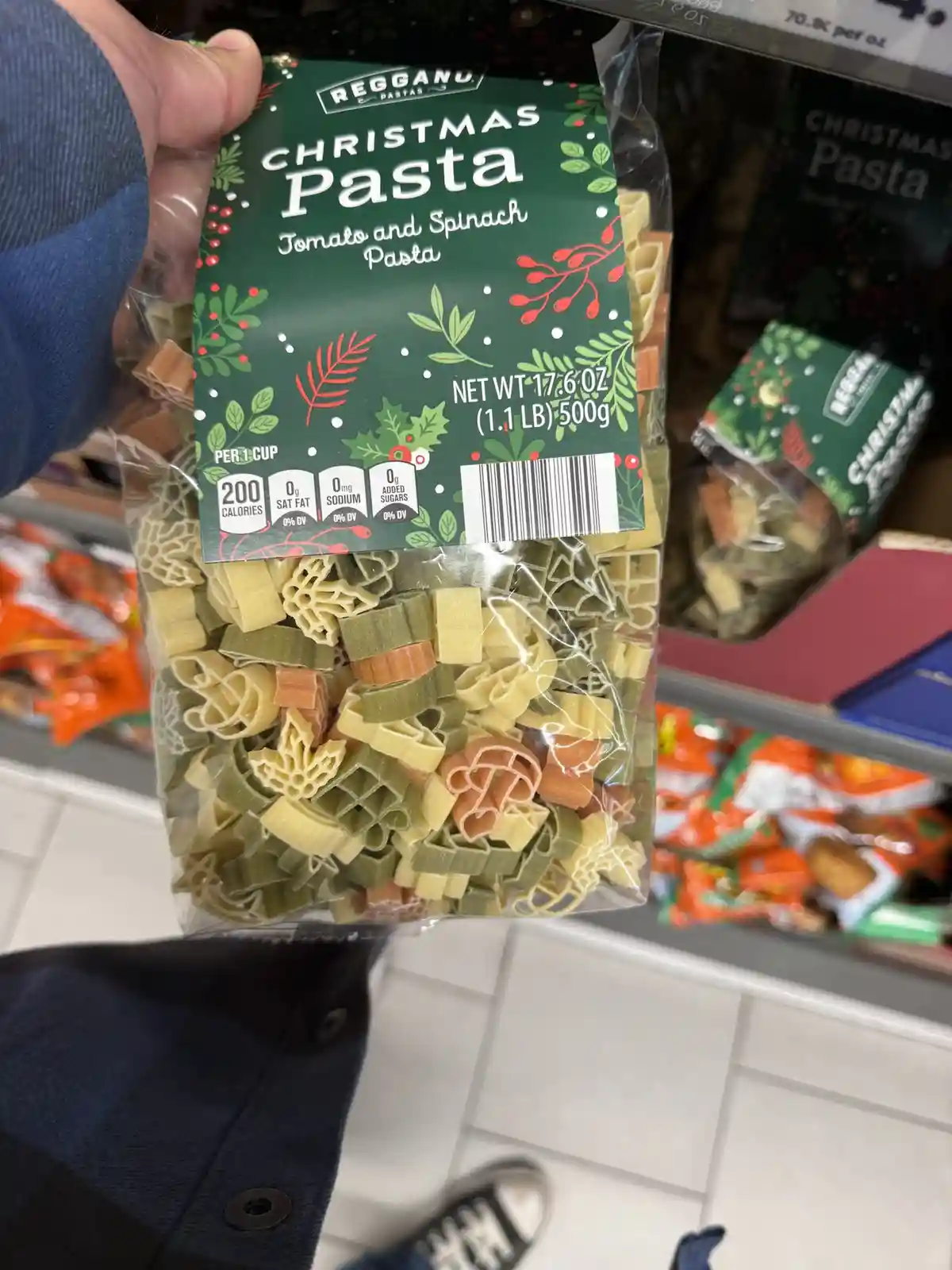 A person holds a bag of Reggano brand 'Christmas Pasta' in a grocery store aisle. The pasta is shaped like holiday ornaments…