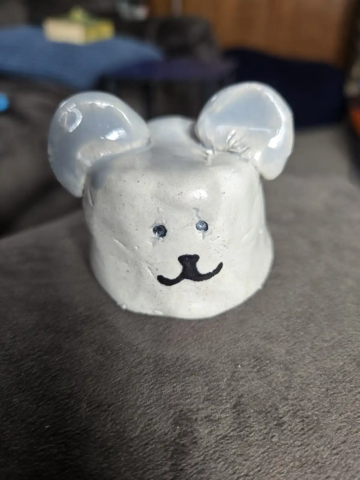A handmade ceramic figurine shaped like a mouse or bear with large rounded ears, simple black marker eyes and nose, and a…