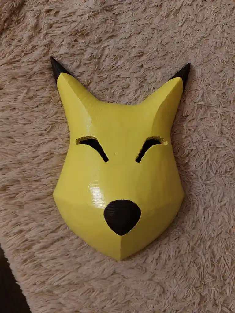 A handmade, yellow, geometrically stylized Pikachu mask with black-tipped ears and simple cutouts for eyes and nose rests on…