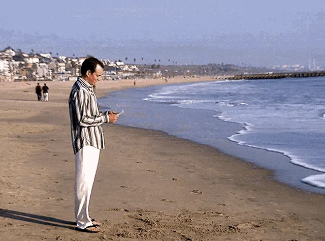 A man in a striped shirt and white pants stands on a beach, throws his phone into the ocean, then turns away as if nothing…